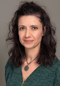 Nő zöld ruhában, arany cipzárral, sötét hullámos hajjal és medállal a nyakában.