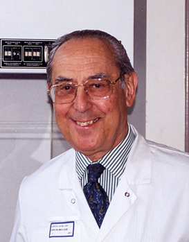 Orvos fehér köpenyben, kék nyakkendővel, orvosi berendezés előtt, jelvénnyel