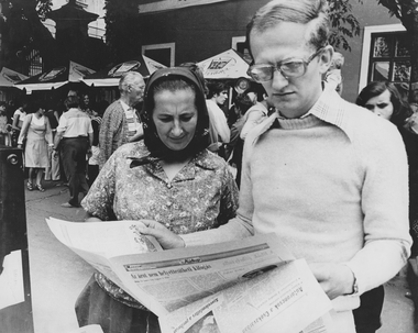 Piac vagy vásár, ahol emberek gyűltek össze. Egy nő és egy férfi újságot olvasnak az előtérben.