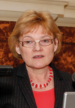 Egy személy piros ruhában és nyaklánccal, mikrofon és laptop mellett látható.