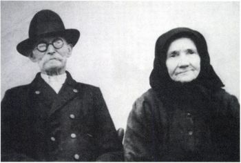 Régi fekete-fehér fotó kalappal és kendővel Régi fekete-fehér fotó, amelyen két ember ül egymás mellett, egyikük kalapot, másik kendőt visel.