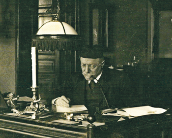 Régi íróasztal, rajta papírok, gyertyatartó és asztali lámpa egy klasszikus belső térben.