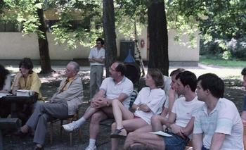 Csoport emberek egy szabadtéri megbeszélésen, fák között ülve.