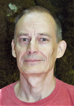 Egy személy piros pólóban, homályosított arccal, zöld növényzet háttérrel.