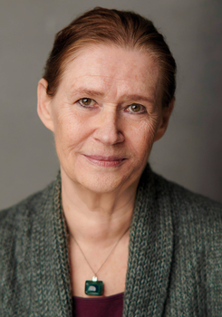 Egy személy szürke kötött kardigánt visel, zöld medálos nyaklánccal.