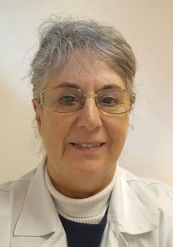 Egy személy fehér orvosi köpenyben, szürke hajjal, háttérben semleges fal.