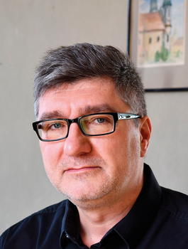 Szürke hajú férfi fekete ingben szemüveggel Szürke hajú férfi fekete ingben szemüveggel, háttérben festmény a falon