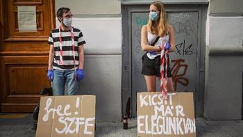 Fiatalok tüntetnek karton táblákkal a SZFE mellett Két fiatal karton táblákkal tüntet, feliratokkal: 'free! SZFE' és 'KICSIT MÁR ELEGÜNK VAN'.