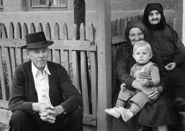 Vidéki környezetben, kerítés mellett ülő idős férfi és két nő, egyikük kisgyermeket tart az ölében.