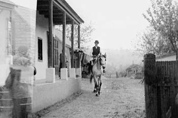 Vidéki ház előtt lovagló férfi Egy vidéki ház előtt lovagló férfi látható, mellette egy elmosódott alak.