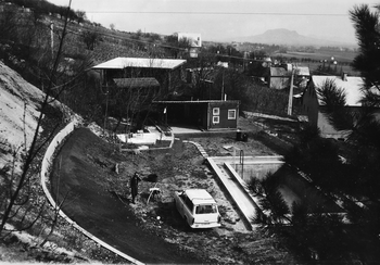 Vidéki ház udvara autóval és kertben dolgozó emberrel Vidéki ház udvara, ahol egy autó parkol és egy ember dolgozik a kertben, háttérben dombok és épületek.