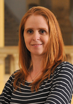 Egy nő vörös hajjal és csíkos felsőben, háttérben homályos épület.